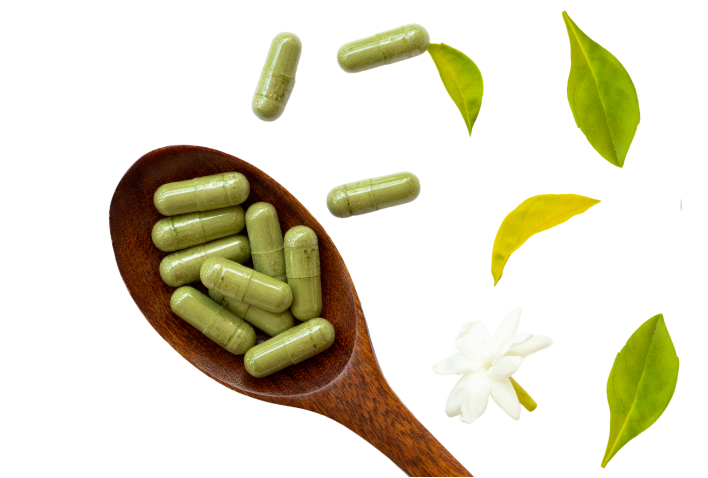 Gruene pflanzliche Kapseln auf einem Holzloeffel mit Blaettern und Blueten. Natuerliche Nahrungsergaenzungen im Feelgood Shop!